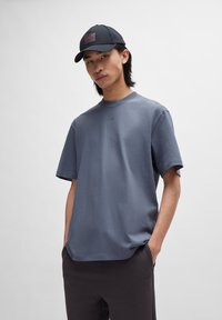 Tricou gri din bumbac cu o croială lejeră și mâneci scurte, având un logo discret al brandului în centru. Sapcă neagră cu un patch roșu al logo-ului.