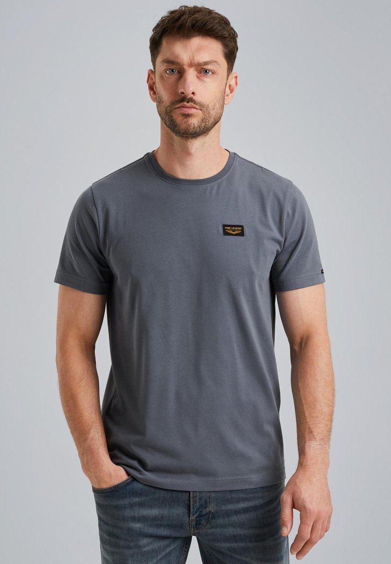 Siva bombažna majica s krožnim izrezom, kratkimi rokavi in črno nalepko na prsih. Gladka tekstura in sproščen kroj.