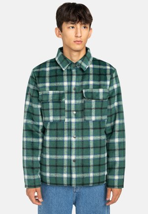 Veste en flanelle écossaise verte avec poches poitrine doubles, devant à boutons et col. Texture douce avec des carreaux contrastés blancs et noirs.