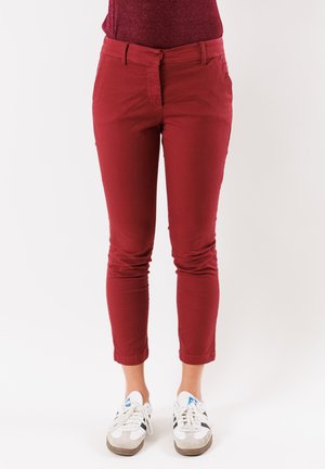 Pantaloni aderenti rossi con un tessuto liscio e vita media. Progettati con due tasche anteriori e una zip. Indossati con sneakers bianche.