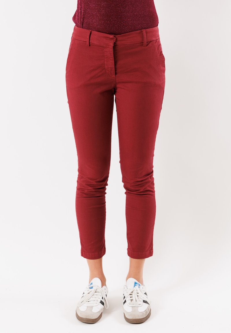Pantaloni aderenti rossi con un tessuto liscio e vita media. Progettati con due tasche anteriori e una zip. Indossati con sneakers bianche.