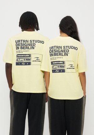 Δύο άτομα φοράνε ανοιχτά κίτρινα T-shirts με μαύρο κείμενο και λογότυπα που γράφουν "URTRN STUDIO DESIGNED IN BERLIN" στην πλάτη, στέκονται δίπλα-δίπλα.