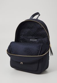 Mochila de nylon azul marino con cremallera dorada, que presenta un forro con estampado y un bolsillo frontal con detalle del logotipo, diseñada para la funcionalidad.