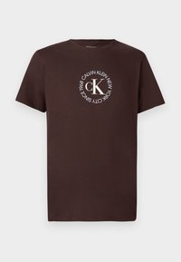 Pruun puuvillane T-särk lühikeste varrukatega, millel on valge ringikujuline logo, mille sees on tekst "CALVIN KLEIN NEW YORK SINCE 1968" ja keskel "cK".