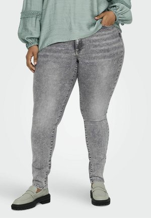 Person trägt eine hellgrüne Bluse, ausgebleichte graue Skinny-Jeans und hellgraue Loafers mit schwarzen Sohlen, steht mit einer Hand in der Tasche.