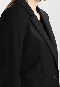 Blazer noir ajusté avec revers cranté, fermeture à bouton unique et coupe structurée. Texture lisse et éléments de design classiques.