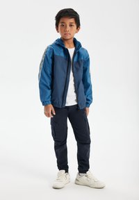 Chaqueta azul con cremallera y capucha, puños elásticos y panel superior de contraste, combinada con pantalones cargo oscuros y zapatillas blancas.