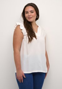 Weiße Bluse mit Rüsche, V-Ausschnitt und strukturiertem Detail am Ausschnitt. Kombiniert mit blauen Jeans. Glatter Stoff.
