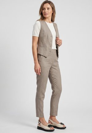 Kvinde iført en beige hørvest og bukser med en hvid kortærmet top og beige slingback-sko, står og kigger sidelæns.