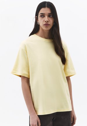 Giovane donna con lunghi capelli scuri e lentiggini che indossa una camicia gialla chiara oversize a maniche corte e pantaloni scuri, guardando leggermente di lato.