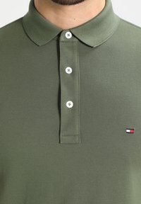 Oliwkowa koszulka polo wykonana z teksturowanej tkaniny. Posiada kołnierzyk, trzy białe guziki oraz małe logo w kolorach czerwonym, białym i niebieskim na piersi.