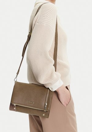 Persona vestida con un suéter de canalé beige y pantalones, con un bolso bandolera de cuero marrón que tiene cierre dorado y detalle de logo.