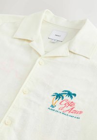 Chemise crème à boutons avec col, palmiers brodés et texte « Côte d'Azur Plage de la Mala, Cap d'Ail » sur la poitrine.