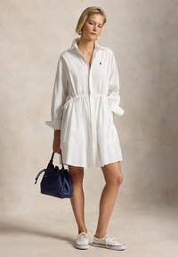 Kvinne iført en hvit kjole med lange ermer, holder en navyblå håndveske og har på seg hvite joggesko, står foran en nøytral bakgrunn.