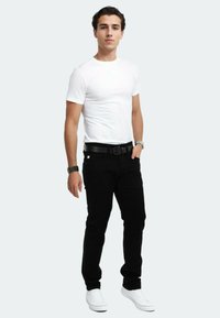 Weißes, eng anliegendes T-Shirt, schwarze Jeans mit silbernen Beschlägen, schwarzer Gürtel und weiße Sneakers. Das Modell steht mit entspannten Händen an den Seiten.