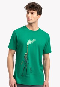 Groene katoenen t-shirt met een grafisch ontwerp van een figuur die een fakkel vasthoudt, omgeven door een speelse rookpatroon. Korte mouwen en een ronde hals.