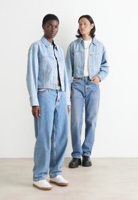 Dos modelos llevan chaquetas de denim azul y pantalones vaqueros holgados. Las chaquetas tienen un patrón texturizado, mientras que los zapatos son zapatillas blancas y botas negras.