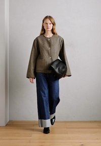 Brun vævet jakke med frontknapper, parret med vide denimbukser og en sort lædertaske; model går på trægulv mod en lys væg.