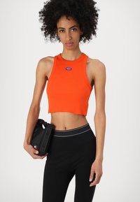 Top court côtelé orange avec un col rond et un patch logo, associé à un legging noir taille haute avec une ceinture de marque.