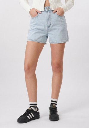 Donna che indossa pantaloncini di jeans azzurri, maglione corto bianco, sneaker Adidas nere e calzini bianchi con strisce nere, in piedi con le mani nelle tasche.