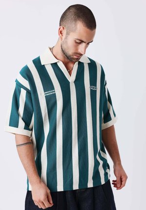 Homme aux cheveux courts et à la barbe portant un polo rayé verticalement turquoise et blanc, regardant vers le bas, mains détendues le long du corps sur fond uni.