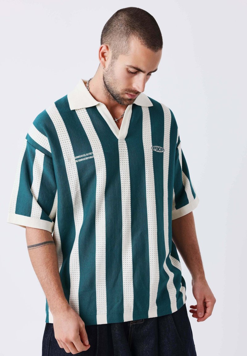 Homme aux cheveux courts et à la barbe portant un polo rayé verticalement turquoise et blanc, regardant vers le bas, mains détendues le long du corps sur fond uni.