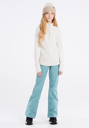 Dames lichte crème top met een ritssluiting, gecombineerd met turkooise skibroek en zwarte sneeuwlaarzen, met een gebreide beige muts.