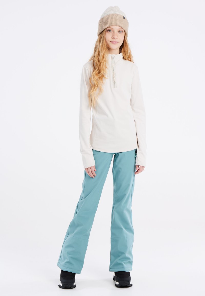Dames lichte crème top met een ritssluiting, gecombineerd met turkooise skibroek en zwarte sneeuwlaarzen, met een gebreide beige muts.