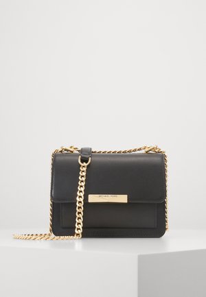 Sac à main en cuir noir Michael Kors avec bandoulière chaîne dorée et plaque nominative rectangulaire dorée sur le rabat avant, posé sur une surface blanche.