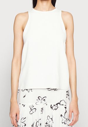 Kobieta ubrana w bezrękawnik w kolorze białym, połączony z białą spódnicą ozdobioną czarnym kwiatowym wzorem, stojąca na jednolitym tle.