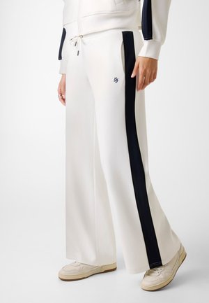 Pantaloni bianchi a gamba larga con strisce laterali nere, tasche frontali e un piccolo logo ricamato vicino alla tasca, indossati con sneakers bianche.