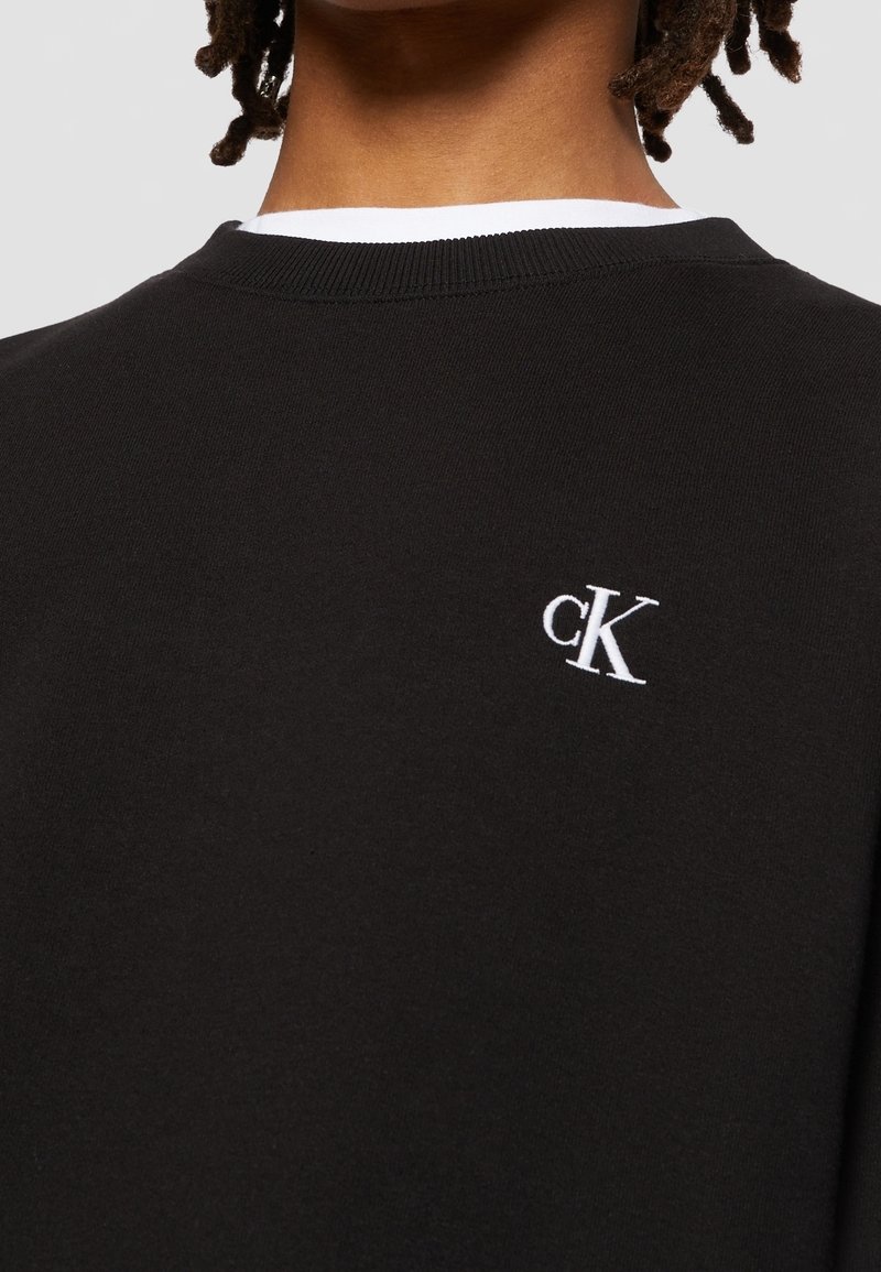Personne portant un pull noir avec un logo blanc "cK" brodé en haut du dos près de l'encolure et une chemise blanche en dessous.