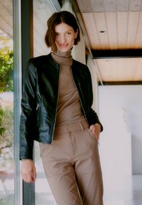 Zwarte leren jas met geribbelde manchetten, tan turtleneck en beige broek. Gladde texturen, op maat gemaakte pasvorm en subtiele details op de kleding.