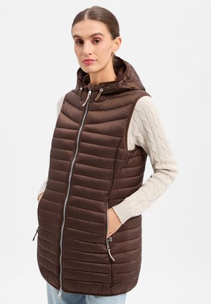 Frau trägt eine braune gesteppte ärmellose Kapuzenweste über einem cremefarbenen Zopfmusterpullover mit den Händen in den Taschen der Weste.