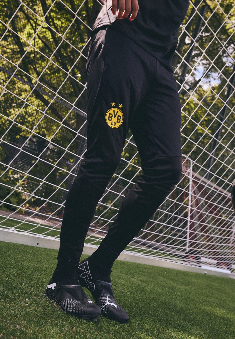 Zwarte sportbroek met een opvallend geel Borussia Dortmund-logo met drie sterren. Draagt zwarte voetbalschoenen en sokken.