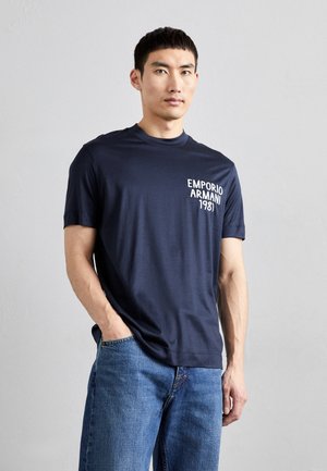 T-shirt blu navy a maniche corte realizzata in tessuto morbido, con un logo e testo bianco sul lato sinistro del petto. Abbinato a jeans blu larghi.