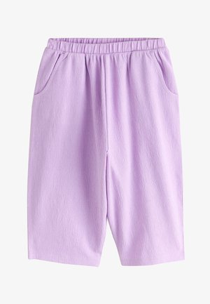 Jong kind staat, glimlacht, draagt een witte mouwloze top met ruches, losse lavendelkleurige broek en witte sandalen met gespen.