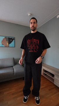 Zwart oversized T-shirt met rood motief aan de voorkant, zwarte wijde broekspijpen en zwarte sneakers met witte veters, staand in een woonkamer.