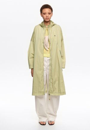 Joven con un abrigo con capucha oversize color verde claro, camiseta amarillo pálido, pantalones blancos de pierna ancha y sandalias negras, posando frente a un fondo blanco.