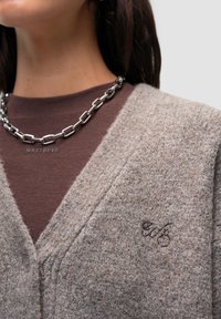 Hnědý vlněný svetr s výstřihem do V, vybavený zapínáním na knoflíky a vyšitým logem. Nosený přes hnědou košili, s stříbrným řetízkem na krku.