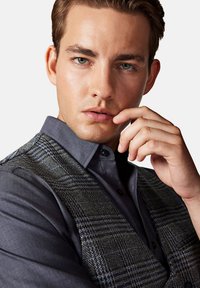 Szary kamizelka w wzory na ciemnej koszuli z guzikami. Kamizelka ma tkany teksturę z geometrycznymi wzorami, a koszula ma gładkie wykończenie.