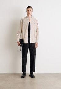 Šviesiai smėlinis marškinėlių mygtukų marškinys, juodas tank top, laisvi juodi kelnių, juodi batai, laikantis mažą juodą krepšį, prieš neutralų foną.
