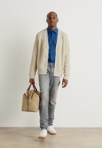 Cárdigan beige de canalé sobre una camisa azul, jeans gris claro y zapatillas blancas. Lleva un bolso marrón claro con un detalle de hebilla.