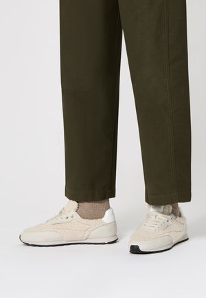Eine Person trägt olivgrüne Hosen, beigefarbene Socken und cremefarbene Sneakers mit Fleecefutter und steht vor einem weißen Hintergrund.