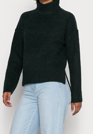 Personne portant un pull à col roulé côtelé noir et un jean taille haute bleu clair, debout devant un fond uni.