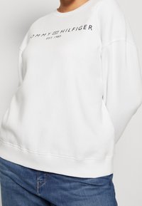 Sudadera blanca de tejido suave, con cuello redondo y hombros caídos. Logo negro "TOMMY HILFIGER EST. 1985" en la parte delantera.