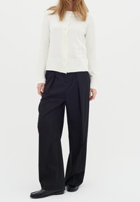 Bílá žebrovaná cardigan s knoflíkovým zapínáním a texturovanými detaily, spárovaná s černými kalhotami s širokými nohavicemi a černými mokasíny.