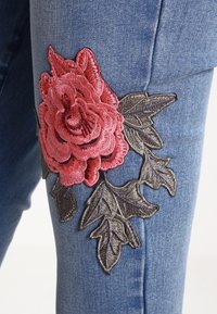 Blå denimjeans med en framträdande broderad ros i rosa och mörkgrön, med detaljerade bladdetaljer, placerade på lårområdet.