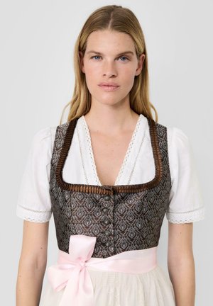 Frau trägt traditionelles braun gemustertes Dirndl-Korsett über weißer Bluse mit Puffärmeln und einer rosa Schleife in der Taille.