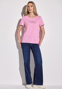 Light pink cotton t-shirt with short sleeves and “NEVER DOUBT yourself” print, paired with dark blue flared jeans and white sneakers.